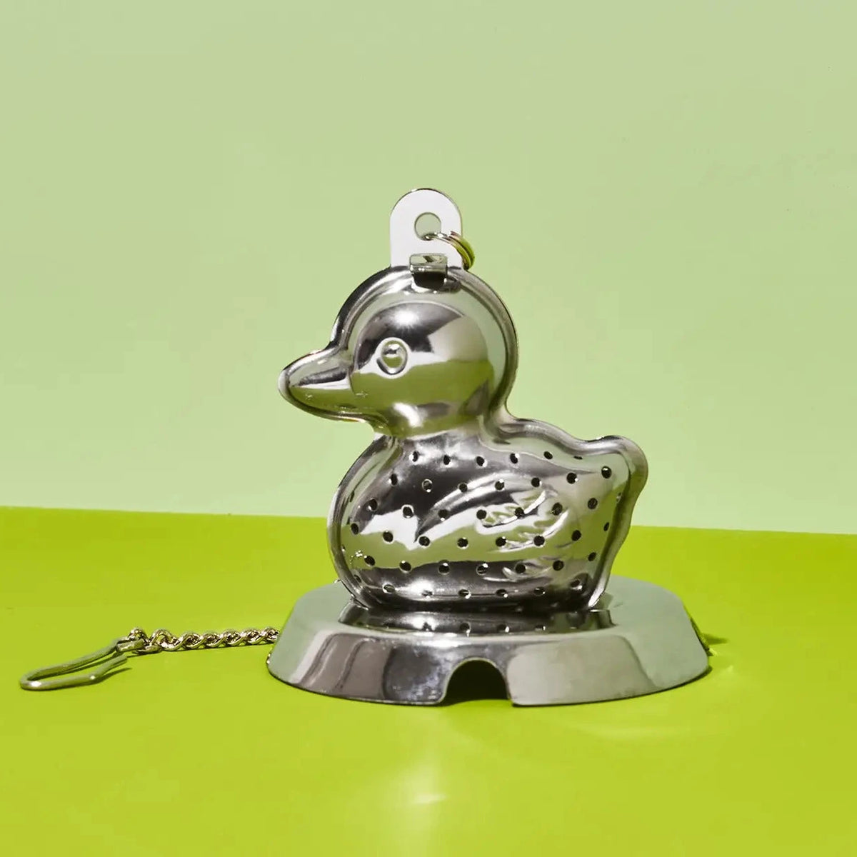 A stainless steel duck-shaped strainer with a small chain attached, shown on a bright yellow-green surface against a matching light green background.