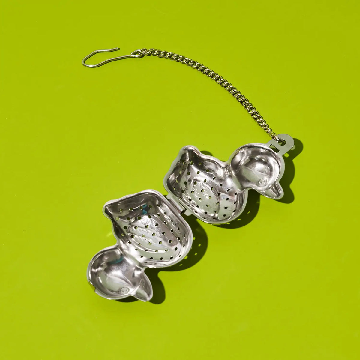 A silver metal tea infuser with a hanging chain and hook, shown open on a bright lime green background.