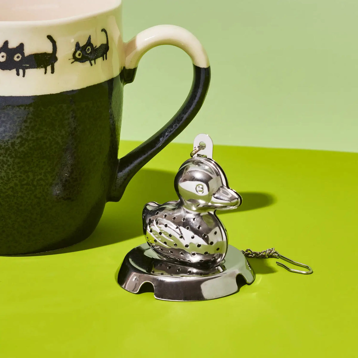 A stainless steel duck-shaped tea infuser with a small hanging key ring, displayed on a bright green surface next to a dark green and cream ceramic mug with black cat illustrations.