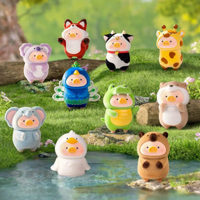 A group of cute animal-shaped duck figurines standing on grass by a small stream in a garden setting. The figures include a fox, cow, giraffe, peacock, frog, bear, duck, and other animal costumes in many colors.
