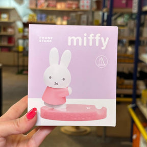 A hand with pink nail polish holds a light pink and white box for a Miffy phone stand in a retail store. The packaging features the character Miffy standing on a pink base, with the text 'PHONE STAND', 'miffy', and a circular logo.