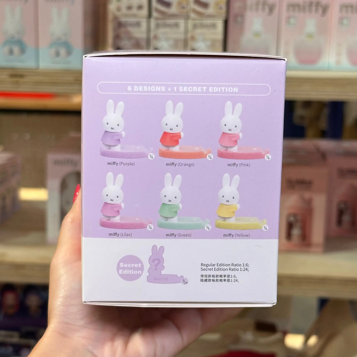 A hand holds a light purple box for a Miffy figurine set in a store. The box shows six Miffy designs in different pastel colors and text reading '6 DESIGNS + 1 SECRET EDITION' and 'Regular Edition Rating 1.