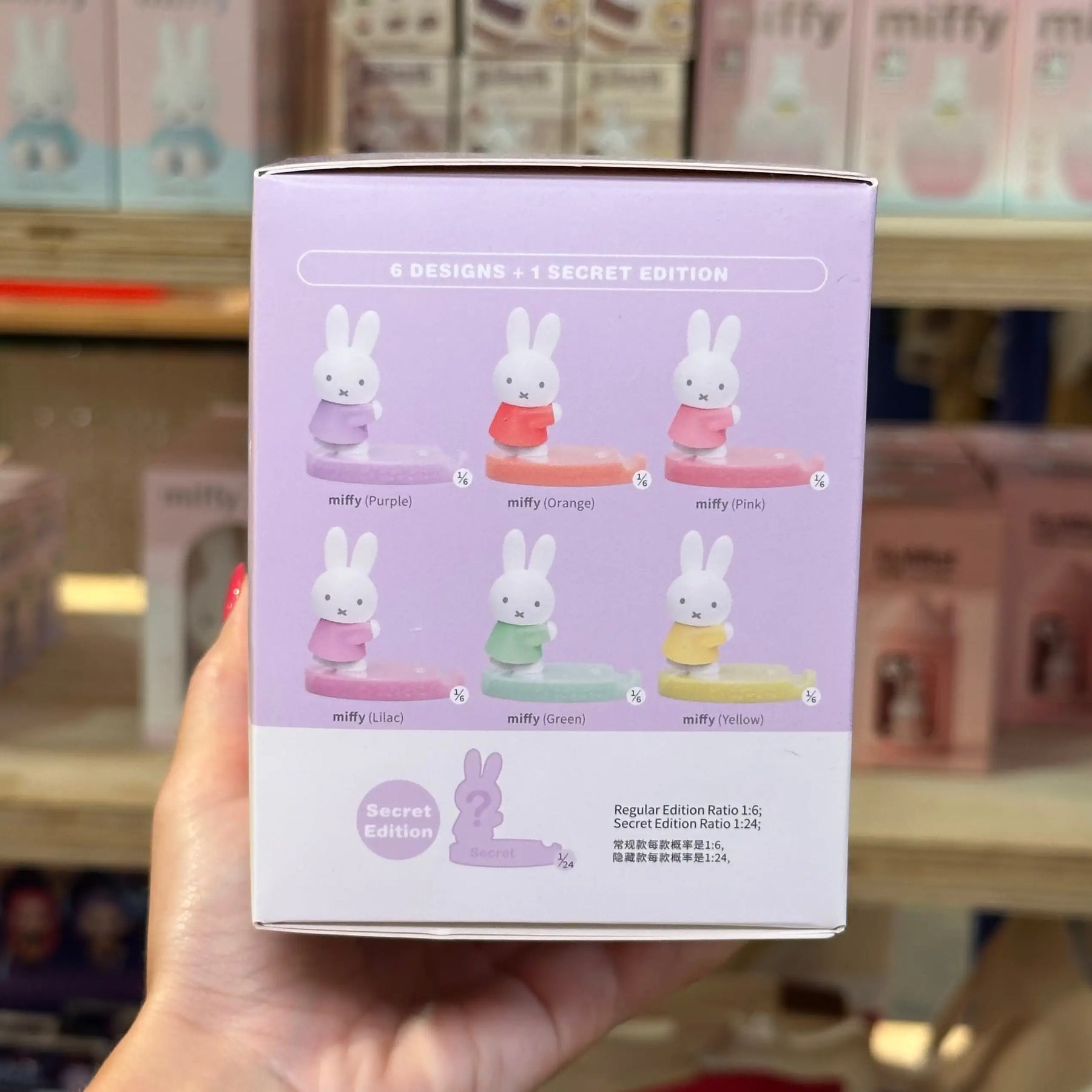 A hand holds a light purple box for a Miffy figurine set in a store. The box shows six Miffy designs in different pastel colors and text reading '6 DESIGNS + 1 SECRET EDITION' and 'Regular Edition Rating 1.