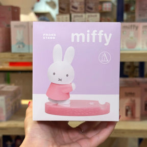 A hand holds a pink and white Miffy phone stand box in a retail store. The packaging features the Miffy rabbit character standing on a pink circular base, with the text 'PHONE STAND' and 'miffy' on a light purple background.