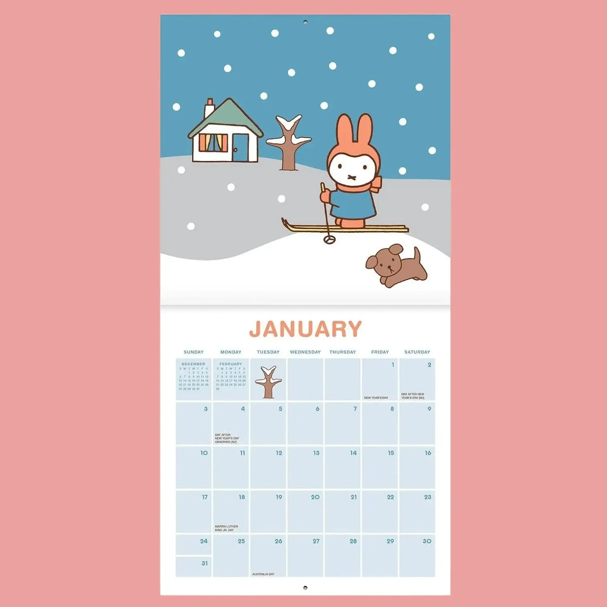 A January wall calendar page featuring a winter illustration with a rabbit skiing, a small house, a tree, and a dog under a blue sky, with a light blue monthly grid below on a pink background.