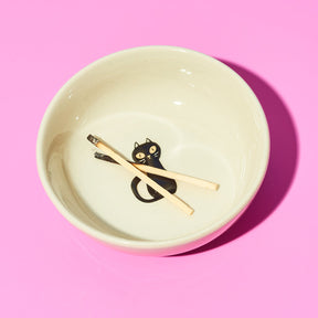 Ceramic bowl with a black cat design and gold utensils on a pink background