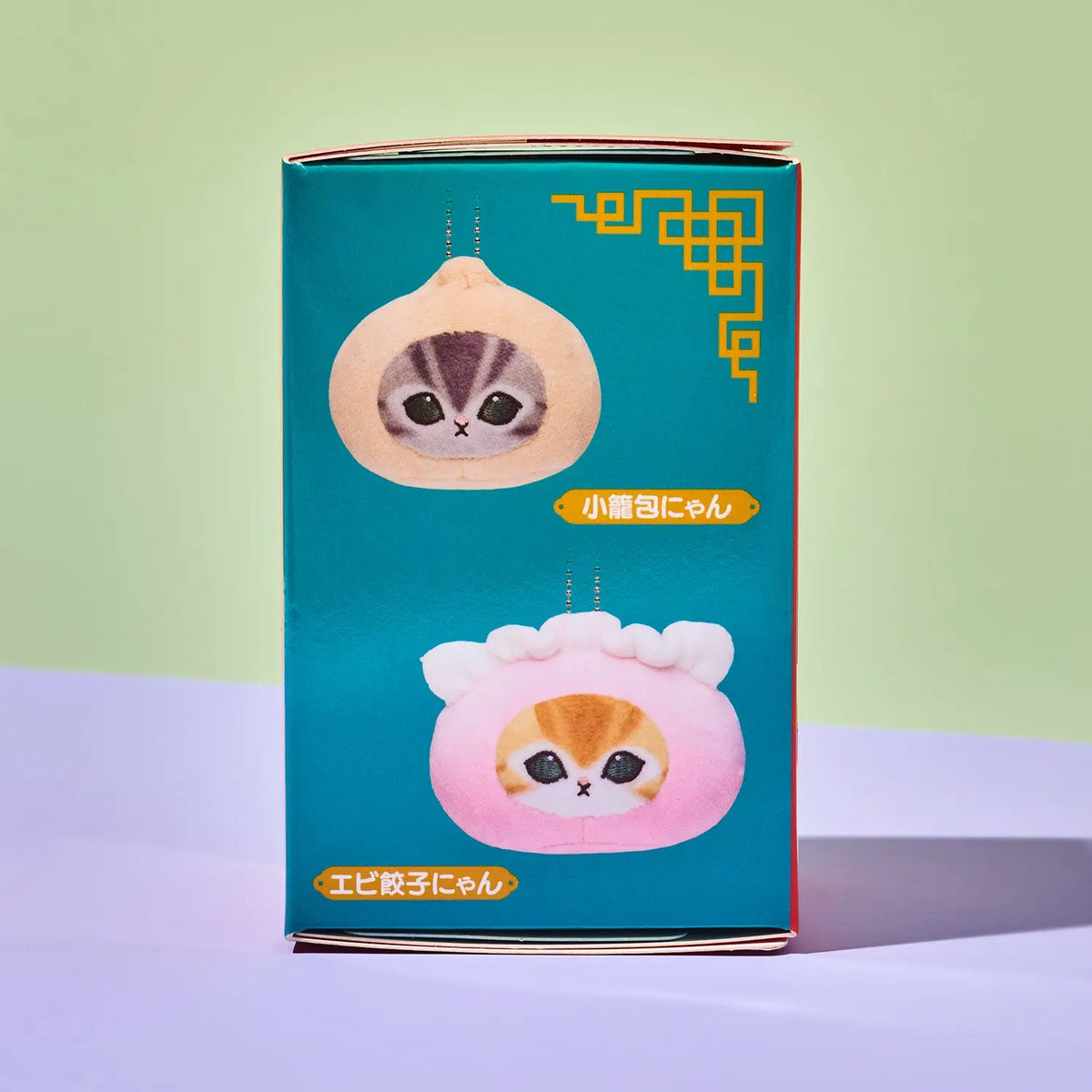 A teal product box with Japanese text and two cute cat-face pouch illustrations, photographed standing on a pale lavender surface against a soft green background.