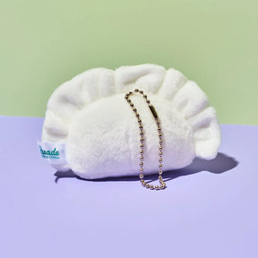 A white plush dumpling-shaped pouch with a gold ball chain strap, resting on a split pastel background of light green and lavender. A white fabric tag on the side reads 'pade with grace'.