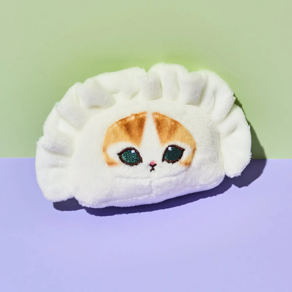 A white plush dumpling-shaped headband or hair tie featuring an embroidered cat face with orange, white, and green details, set against a split green and purple background.