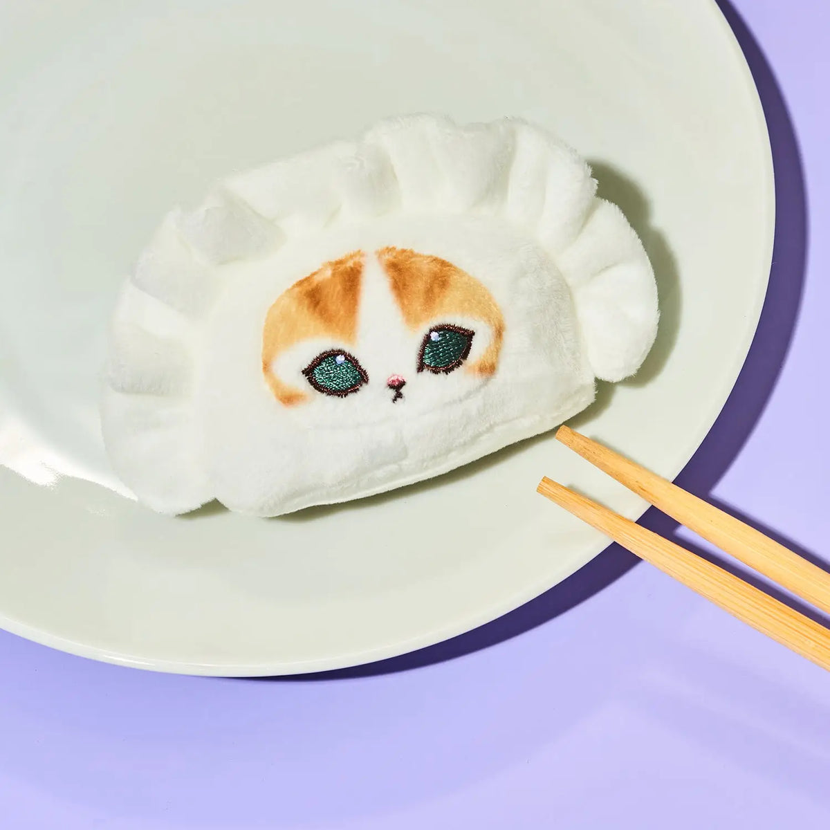 A white plush dumpling-shaped cushion with a cute calico cat face design, resting on a white plate with wooden chopsticks on a light purple background.