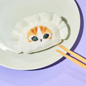A white plush dumpling-shaped cushion with a cute calico cat face design, resting on a white plate with wooden chopsticks on a light purple background.