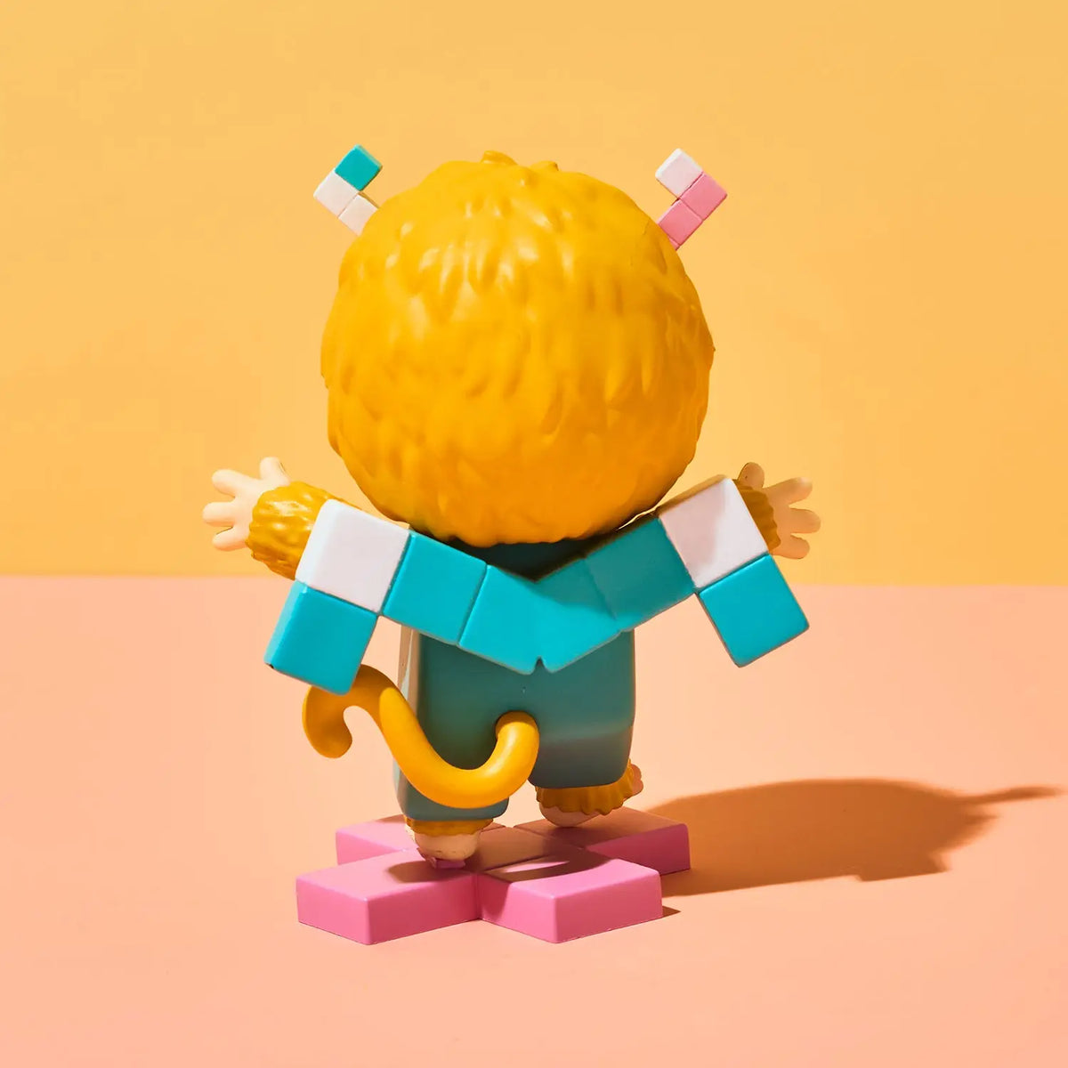 A stylized toy figurine shown from the back, featuring a large yellow textured head, turquoise and white blocky arms, teal body, yellow tail, and pink base, photographed against a warm yellow and peach studio background.
