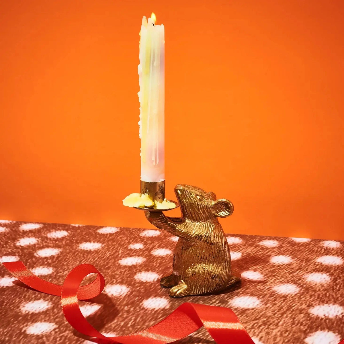 A gold mouse-shaped candle holder with a lit white taper candle, set on a brown and white polka-dotted surface with a red ribbon against a solid orange background.