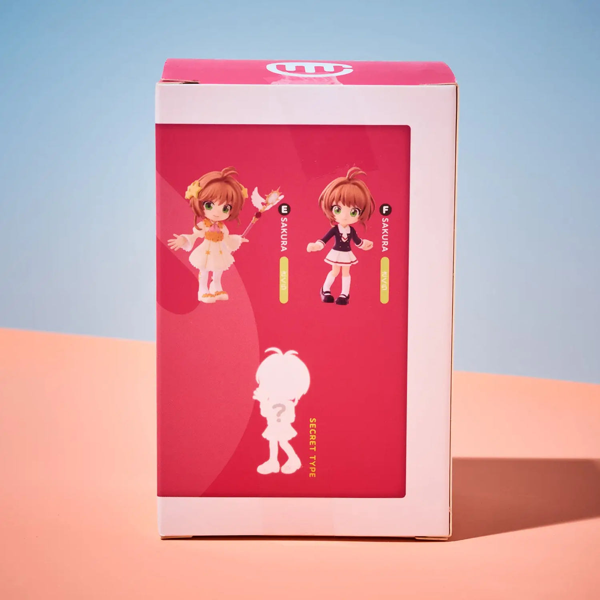 A pink and white retail box featuring anime-style character artwork on the front, photographed standing upright against a soft blue-to-peach gradient studio background.