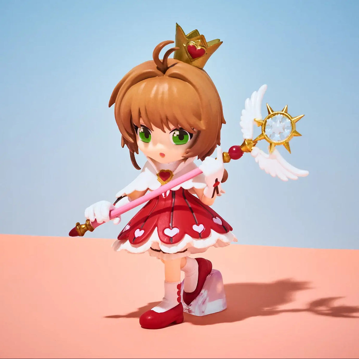 A chibi-style anime figurine of a girl with brown hair and green eyes, wearing a red and white dress with heart details and a gold crown, holding a golden wand with white wings, standing on a peach-colored surface against a light blue background.