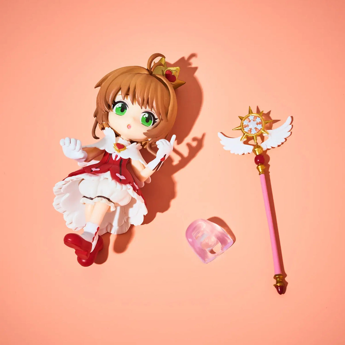 Chibi-style anime figurine of a young girl with brown hair and green eyes, wearing a red and white dress, shown with a pink and gold wand and a small translucent pink accessory on a peach background.