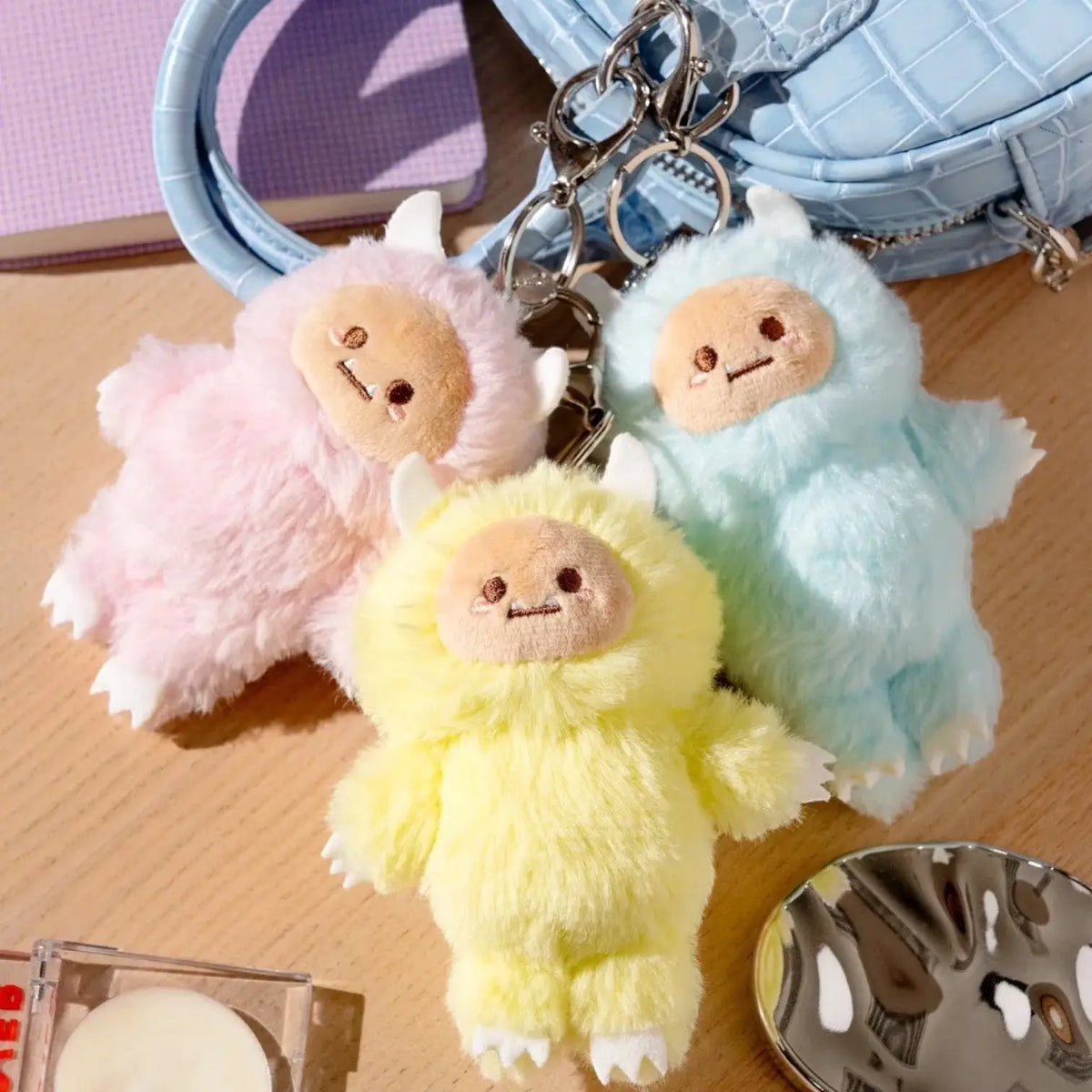 Three colorful fluffy keychains on a wooden surface with a blue textured bag in the background.