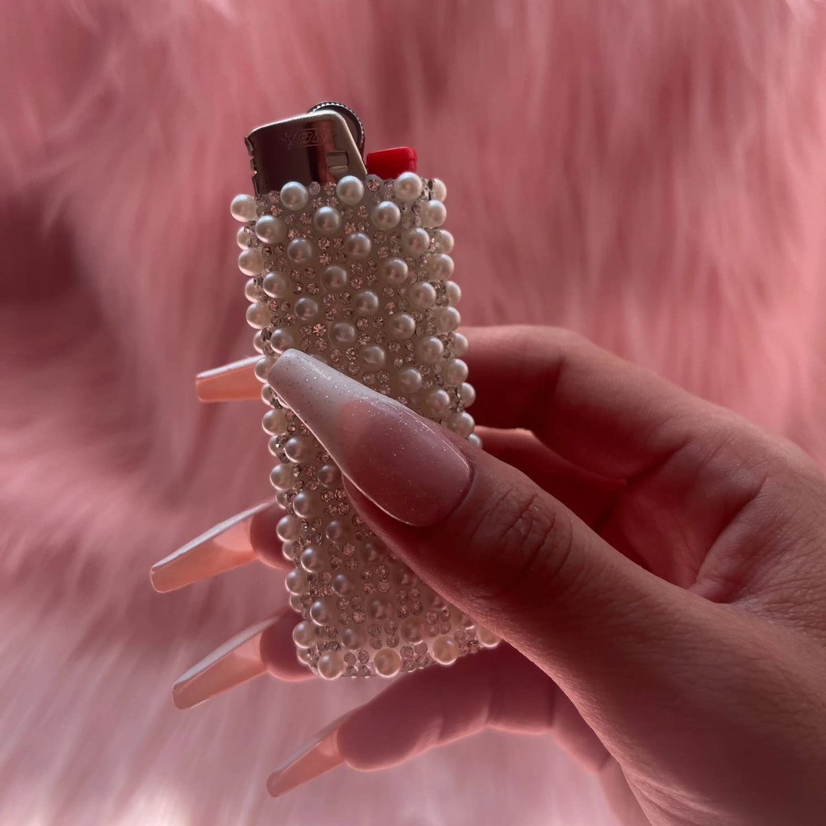 A hand with long nude manicured nails holds a silver lighter wrapped in a lighter covered in white faux pearls, set against a soft pink furry background.