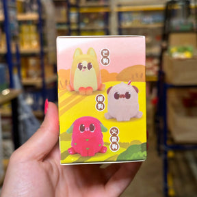 A hand holds a small illustrated product box featuring three cute cartoon dog-like characters in yellow, pink, and light gray, with Japanese text. The background shows a blurred retail or warehouse shelf environment.
