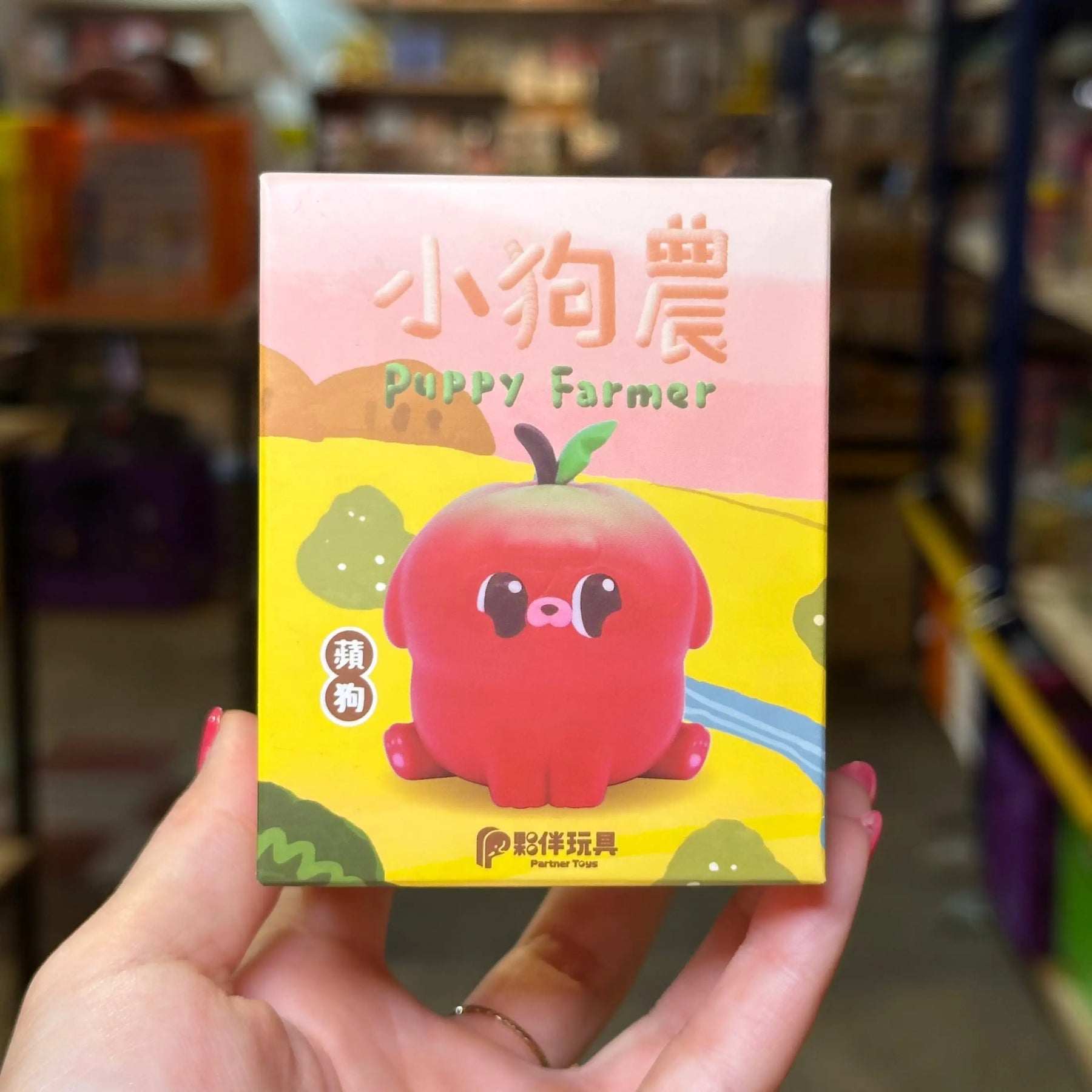 A hand holds a colorful product box labeled 'Puppy Farmer' with a cartoon red puppy character on the front, photographed in a blurred store or shop interior background.