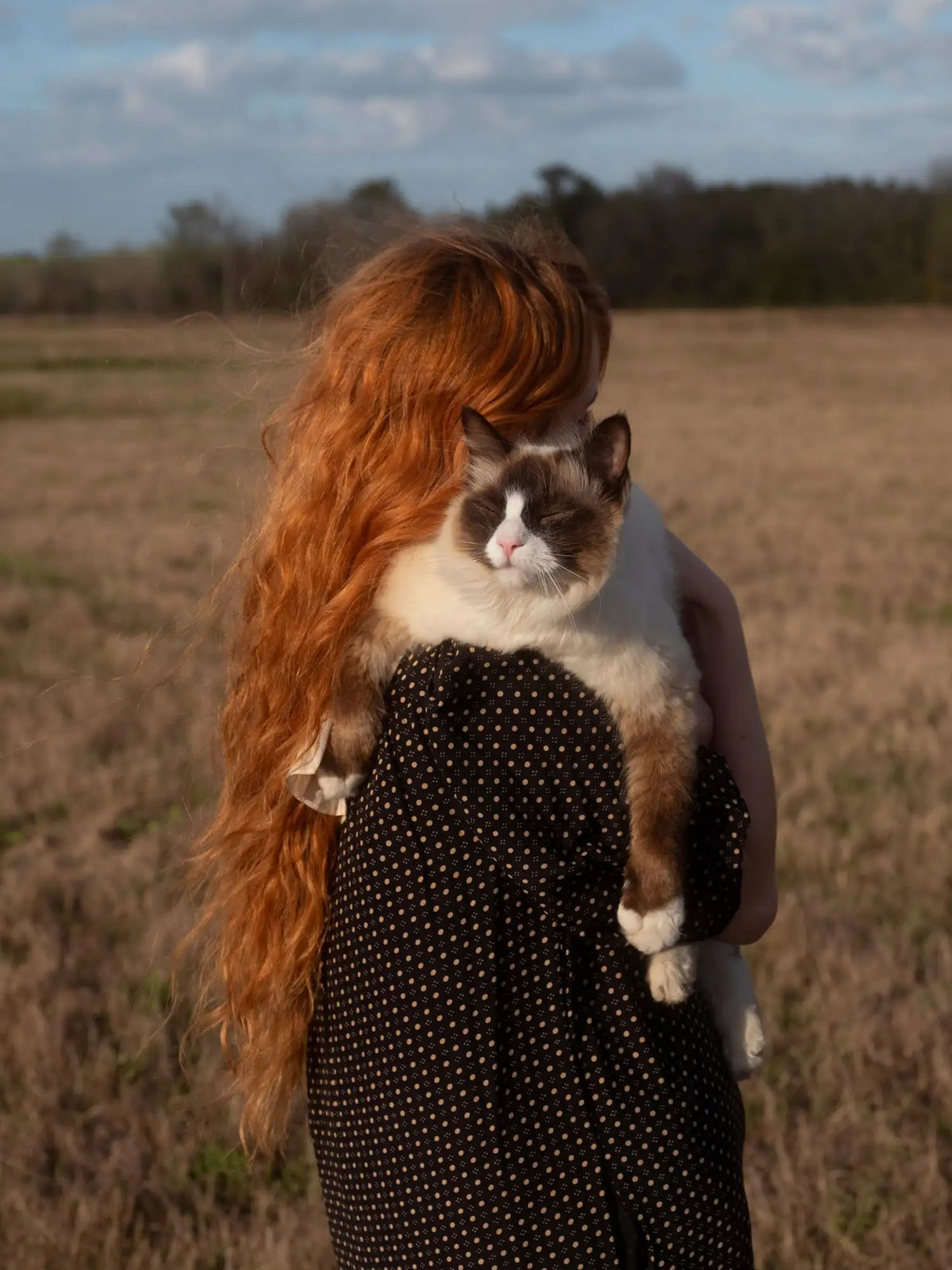 Woman holding a cat in an open field with a cloudy sky