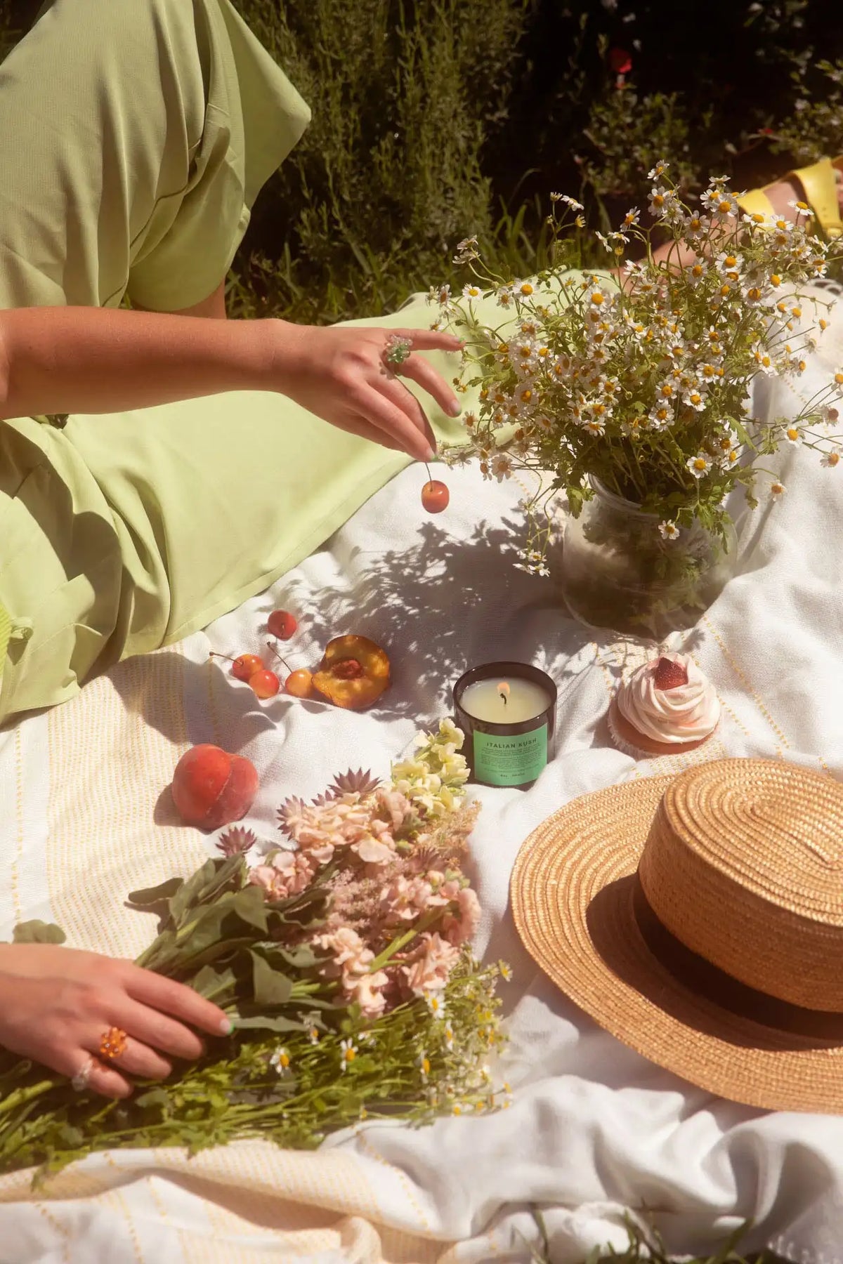 Person arranging flowers on a picnic blanket with a straw hat and snacks.