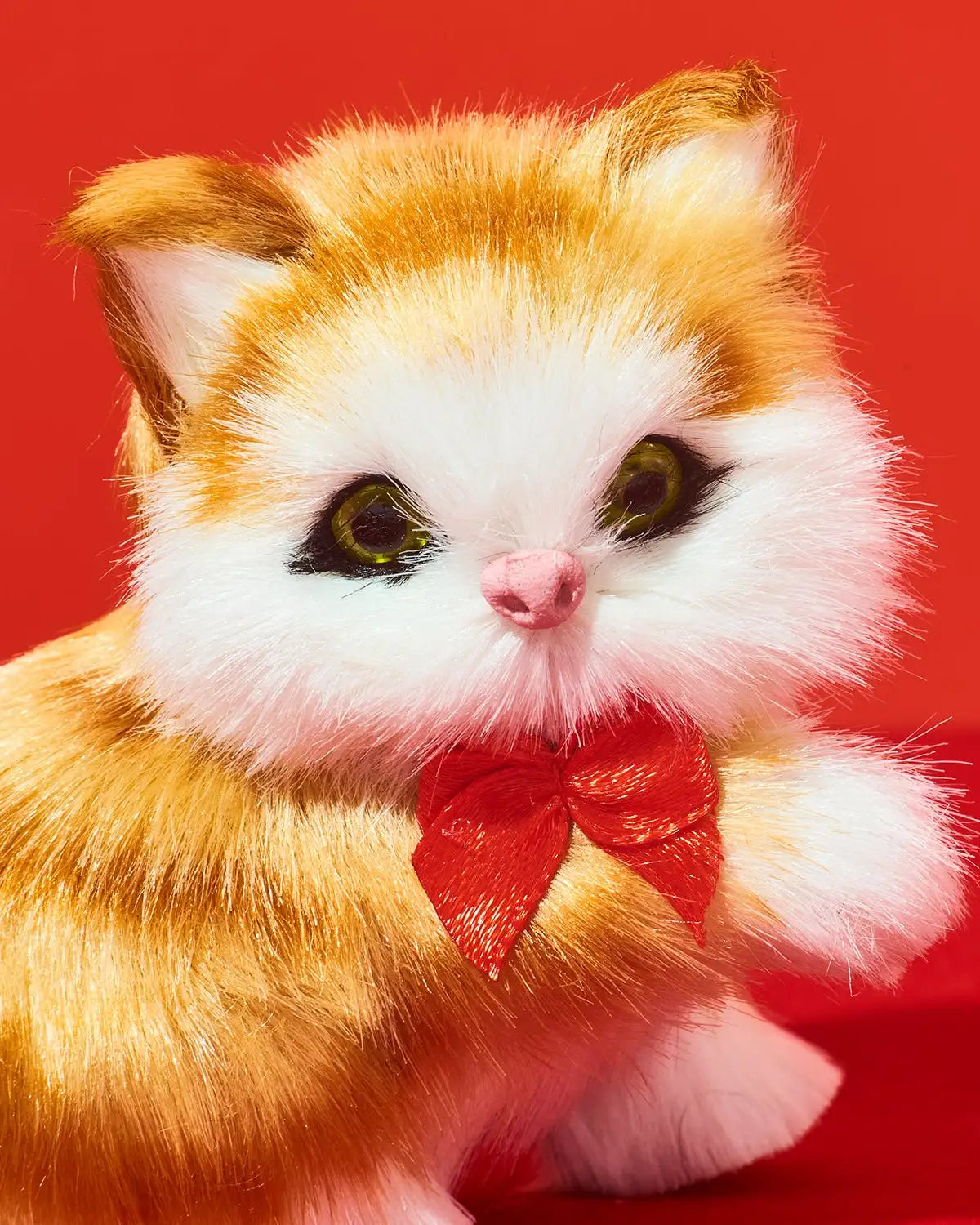 A plush orange-and-white kitten toy with large green eyes and a red bow around its neck, posed against a solid red background.