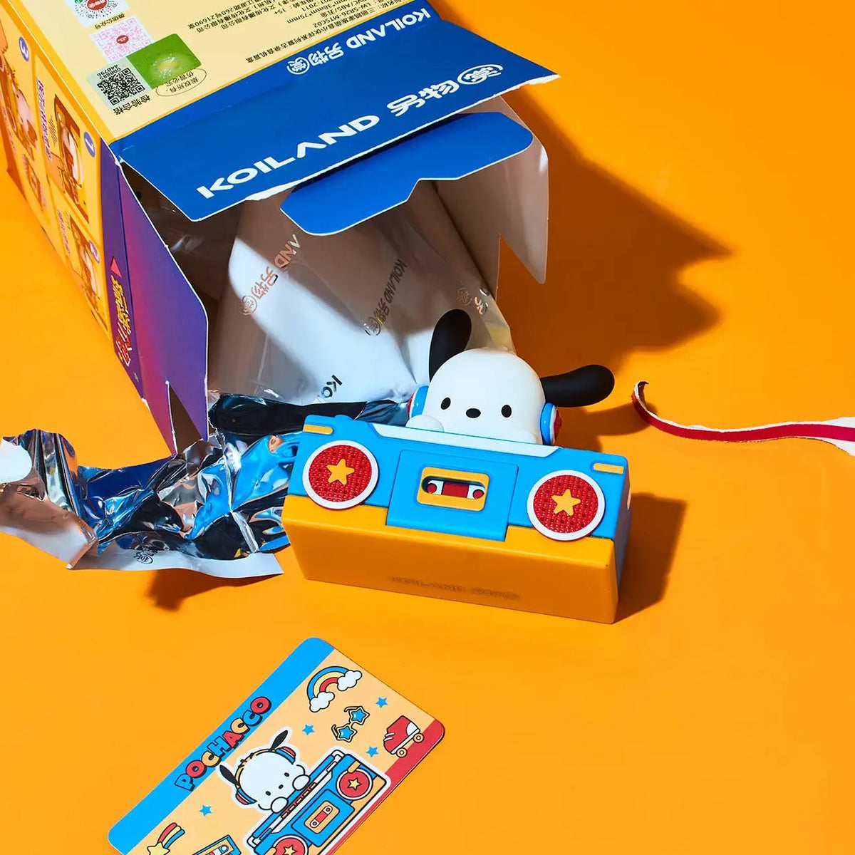 A colorful toy cassette player shaped like a cartoon dog sitting in a cassette car, shown with its blue and yellow packaging box, a small card, and a foil wrapper on a bright orange background. The box shows the brand name KOILAND.