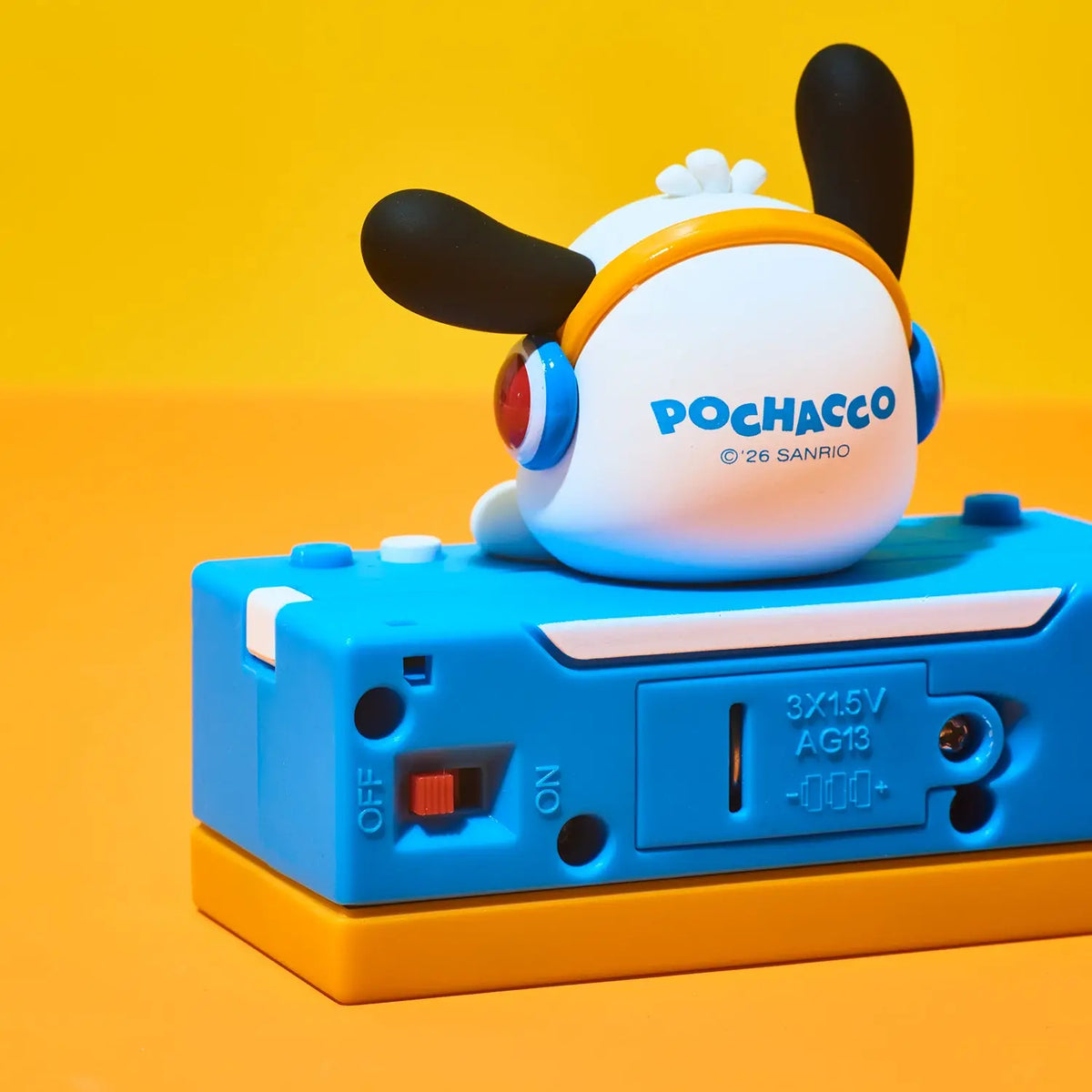 A Pochacco toy figure wearing blue headphones sits on top of a blue and orange electronic device against a bright yellow and orange studio background. The toy shows the back side with the Pochacco logo and Sanrio copyright text.