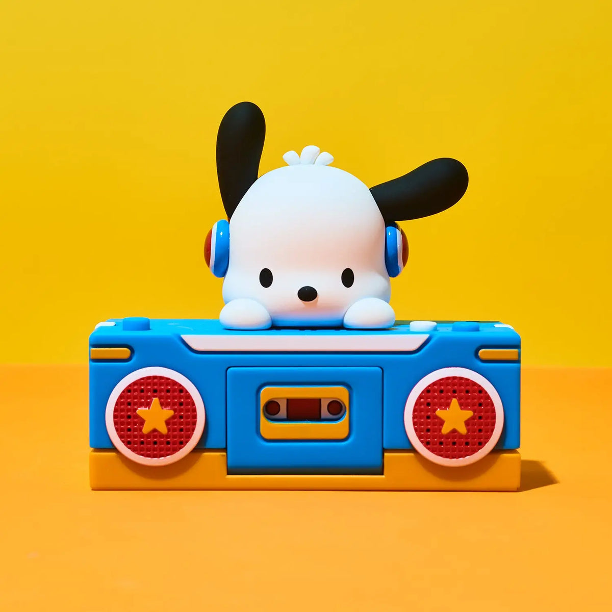 A stylized white cartoon dog character with black ears sits on top of a bright blue cassette-deck-like toy with red circular speaker details, set against a solid yellow and orange background.