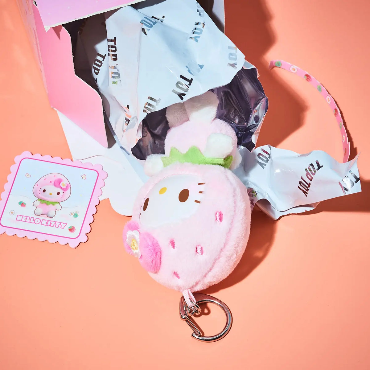 A pink Hello Kitty strawberry plush keychain charm with a metal key ring, shown next to its branded blind bag packaging and a decorative sticker on a peach-colored background.