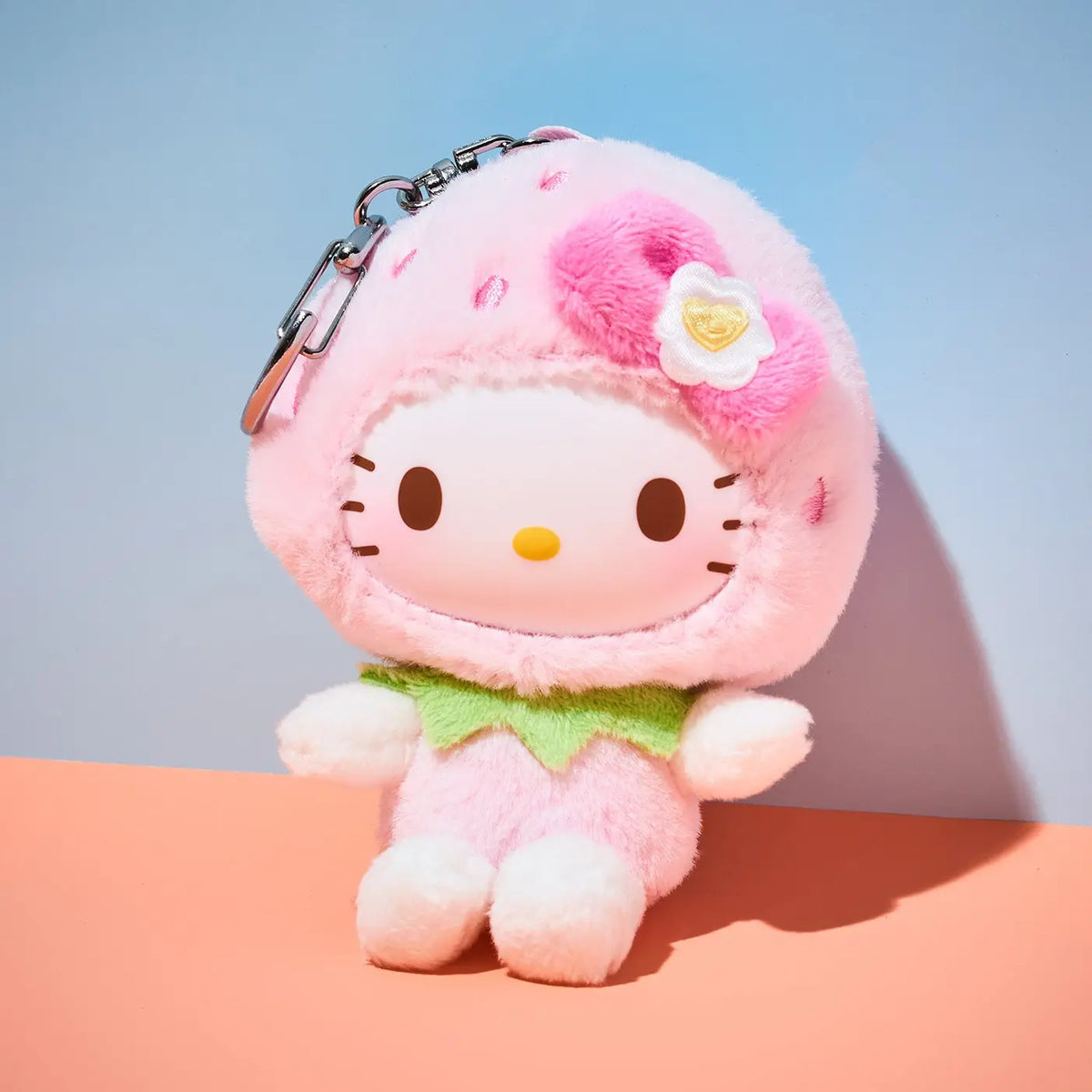 A Hello Kitty plush keychain styled as a strawberry character, wearing a pink strawberry costume with a green leaf collar and a pink flower hair accessory, sitting on a peach-colored surface against a blue background.
