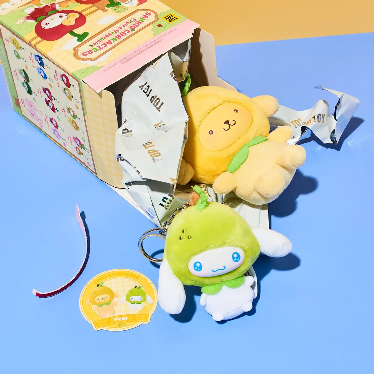 A Sanrio 'Topo de Cartón' blind box toy set shown with two plush keychains: a yellow lion cub and a green pear character with white wings, alongside a branded sticker and packaging on a blue and yellow background.