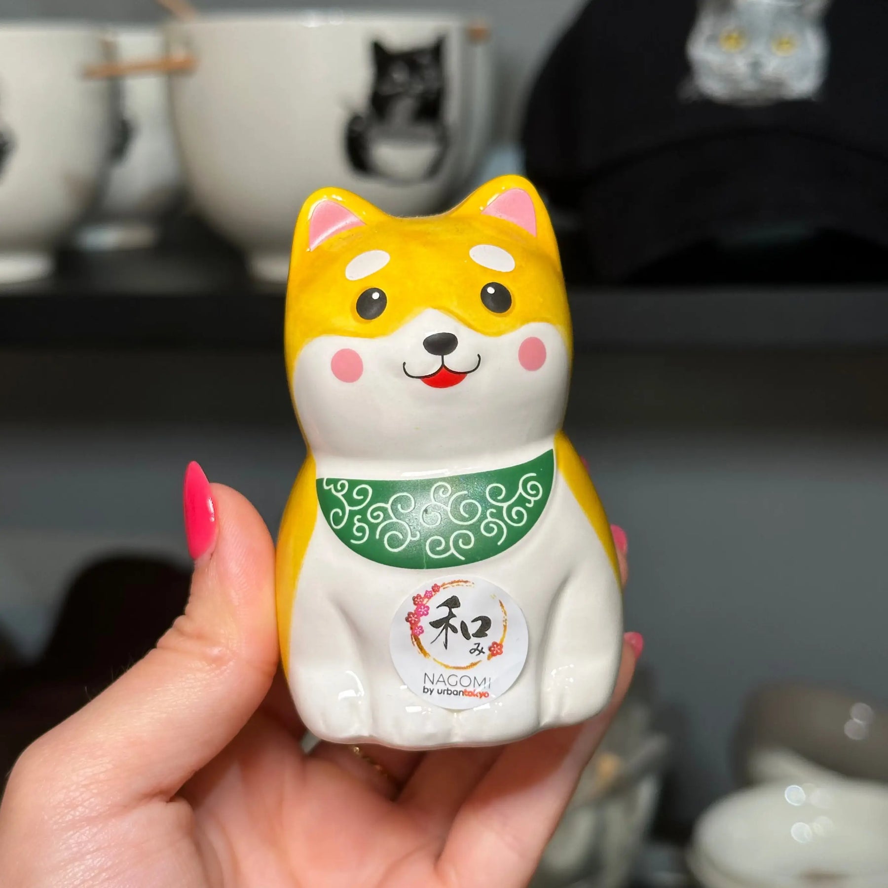 A hand holding a small ceramic Shiba Inu figurine with yellow and white coloring, a green patterned collar, and a white belly with a circular Japanese character emblem. The background shows shelves with white bowls and a black cap.