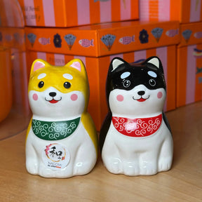 Two ceramic Shiba Inu salt and pepper shakers shaped like yellow, white, black, and white dogs, wearing green and red bands, displayed on a wooden surface with orange boxes in the background.
