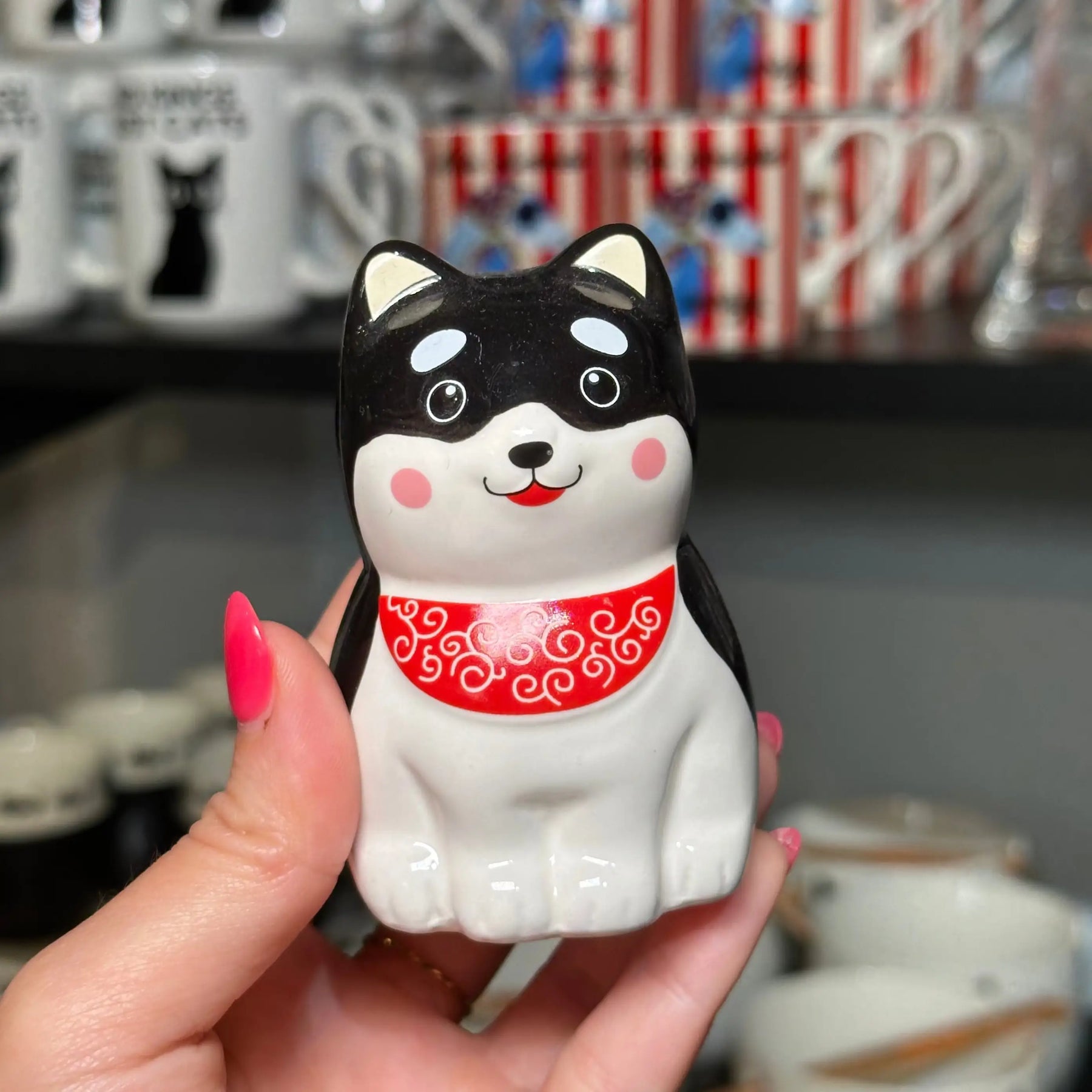 A hand with pink nail polish holds a glossy ceramic cat figurine with a black and white body and a red bandana. The background shows shelves with other mugs and ceramic items in a shop setting.