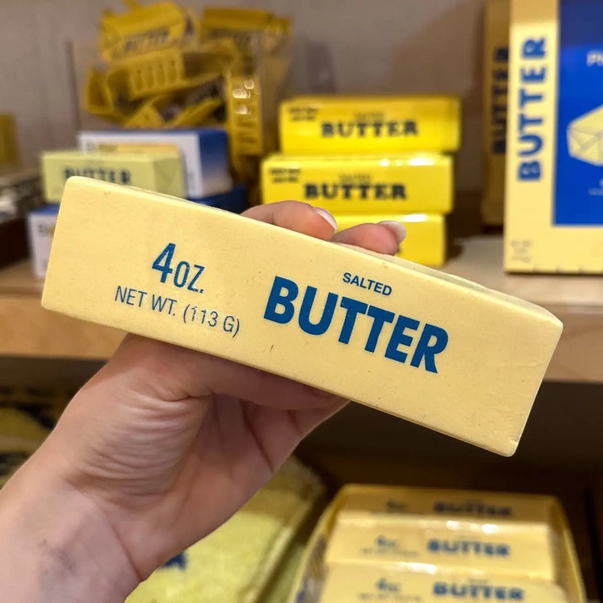 Hand holding a stress toy that looks like a 4oz block of salted butter with more butter products in the background.