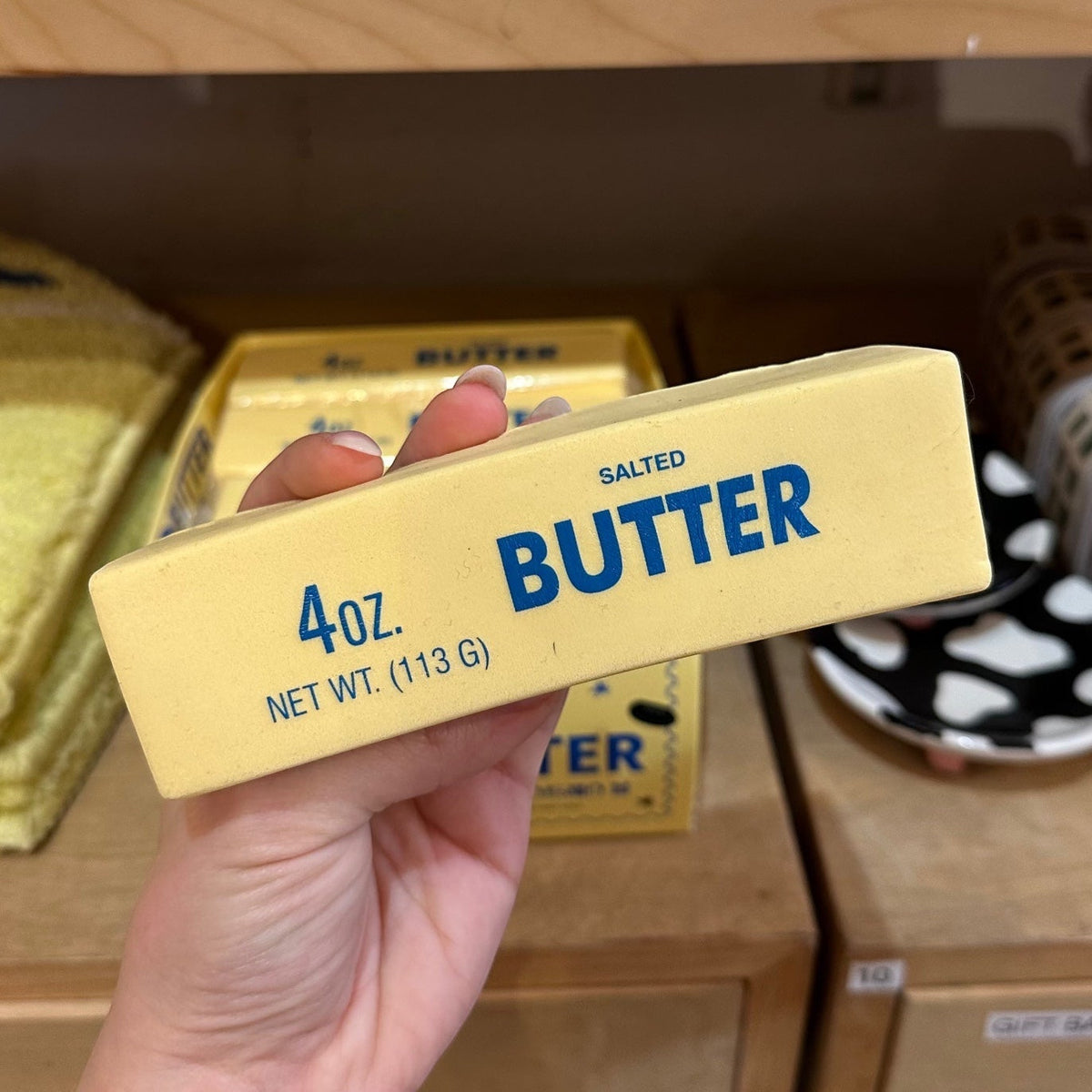 Hand holding a package of salted butter stress toy with a wooden shelf in the background