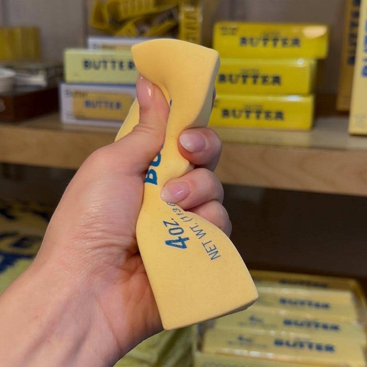 Hand  squishing a salted butter stress toy in front of shelves with similar products.