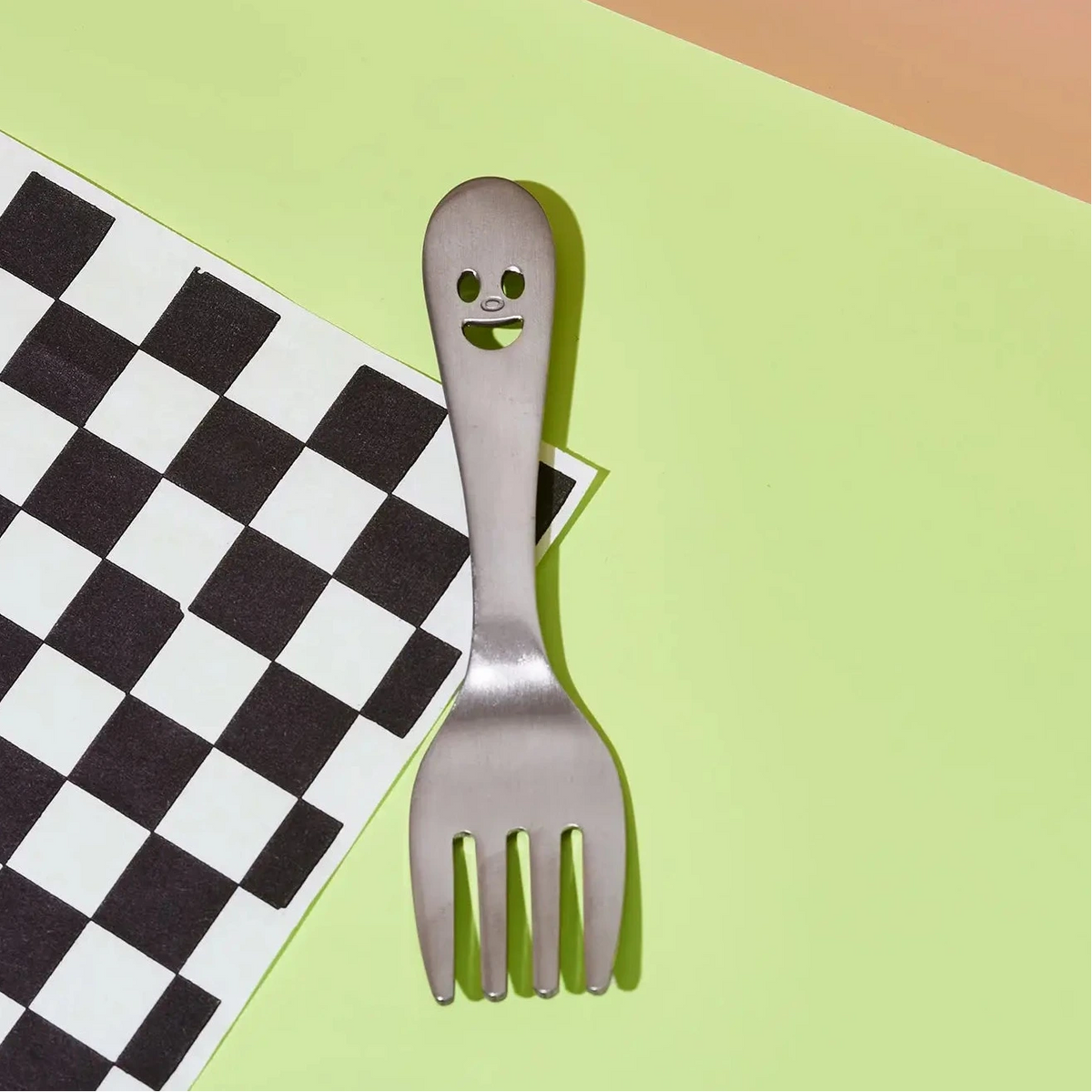 A stainless steel spork-style utensil with a ghost face cutout on the handle, placed diagonally on a black-and-white checkered paper and a light green background.