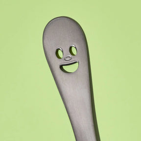 A close-up of a stainless steel utensil handle with a novelty face cutout, set against a solid light green background.
