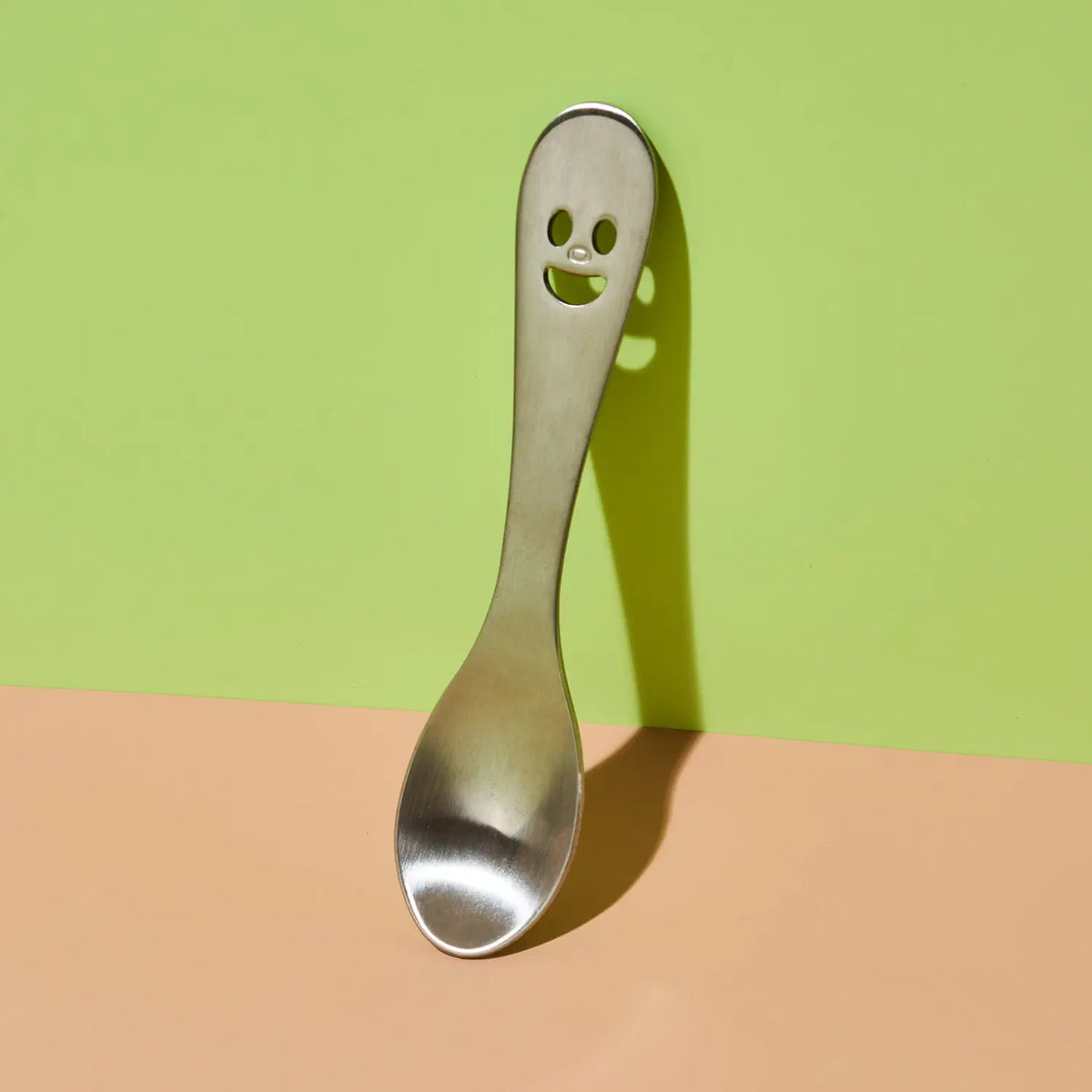 A stainless steel spoon with a ghost face cutout in the handle, standing upright against a split green and peach background.