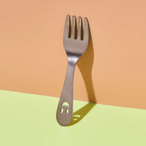 A stainless steel spork with a small smiley face cutout on the handle, standing upright against a two-tone peach and light green background.