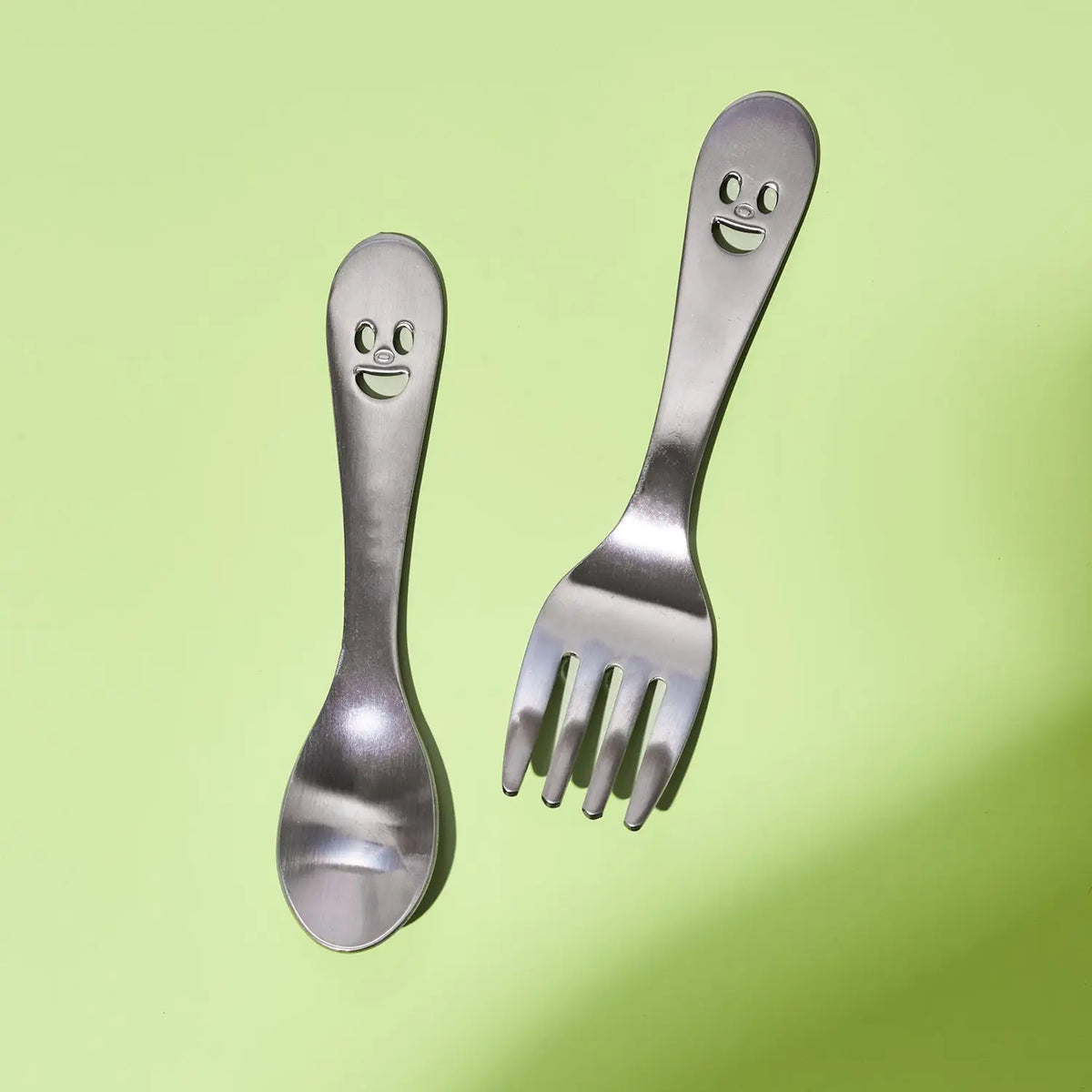A stainless steel children's spork and fork set with smiling face cutouts on the handles, shown on a light green background.