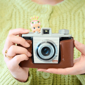 Person holding a vintage-style camera with a small figurine on top, wearing a green sweater.
