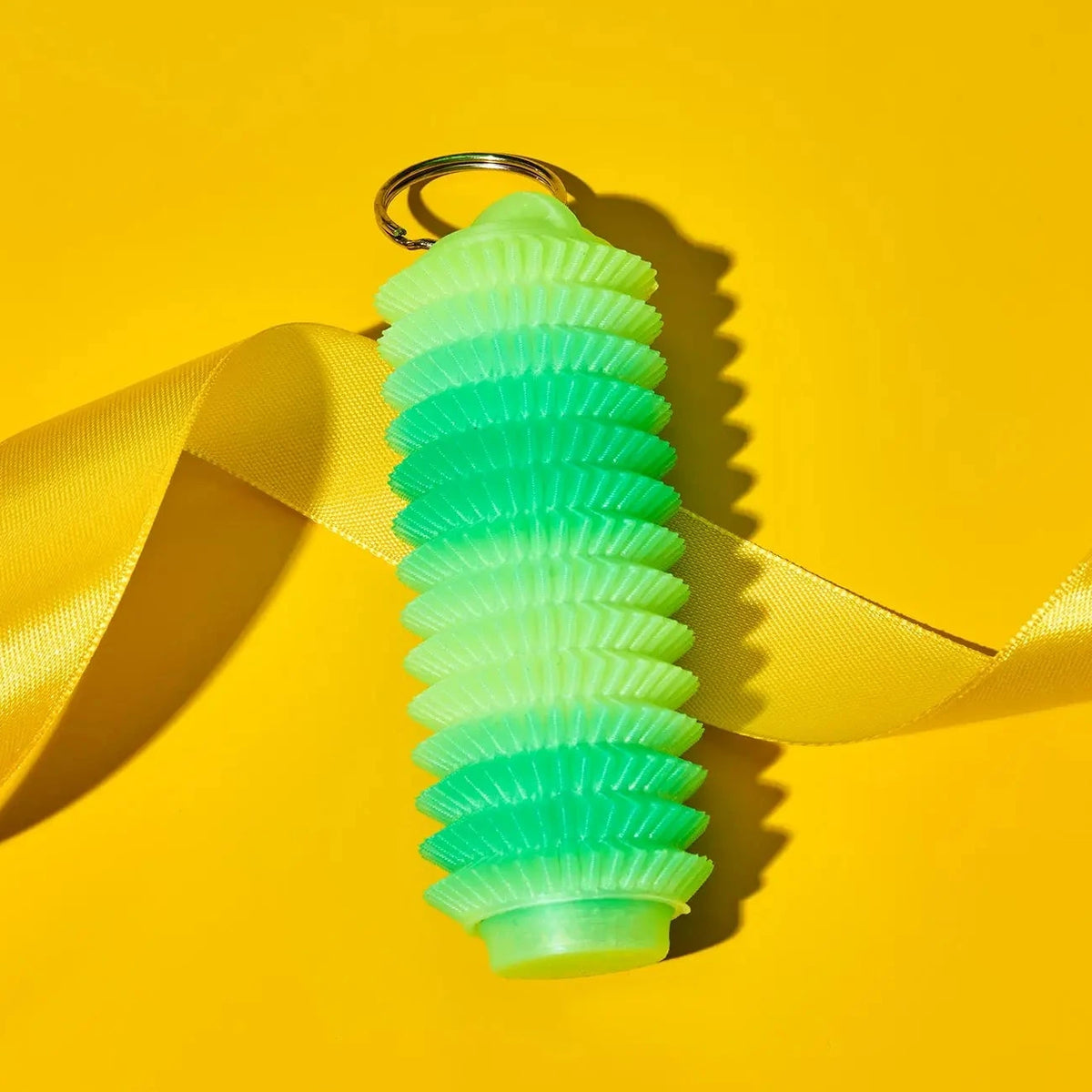 A green gradient fidget keychain holder with a gold metal keyring, wrapped in a yellow satin ribbon against a solid yellow background.