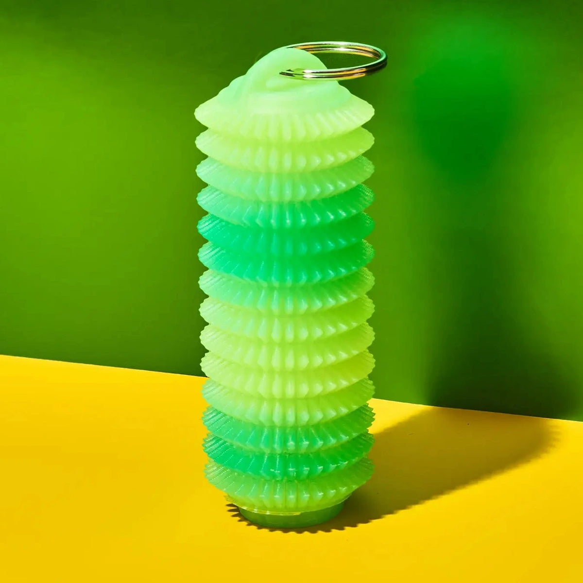 A translucent silicone keychain container shaped like a coiled spring, featuring a green-to-yellow gradient and a silver metal keyring at the top, set against a vibrant green and yellow background.