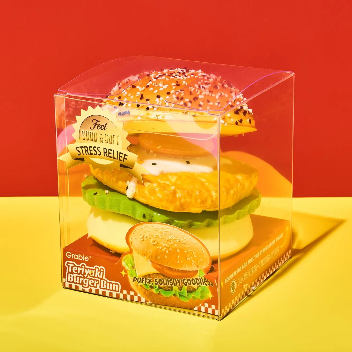 A Grabbii Teryuki Burger Bun stress relief toy inside a clear plastic packaging box, set against a vibrant red and yellow background.
