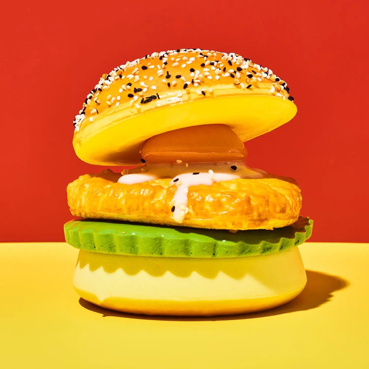 A novelty toy hamburger with a yellow body, sesame seed bun, fried patty, white sauce, green pickle slice, and bottom bun, set against a split red and yellow background.