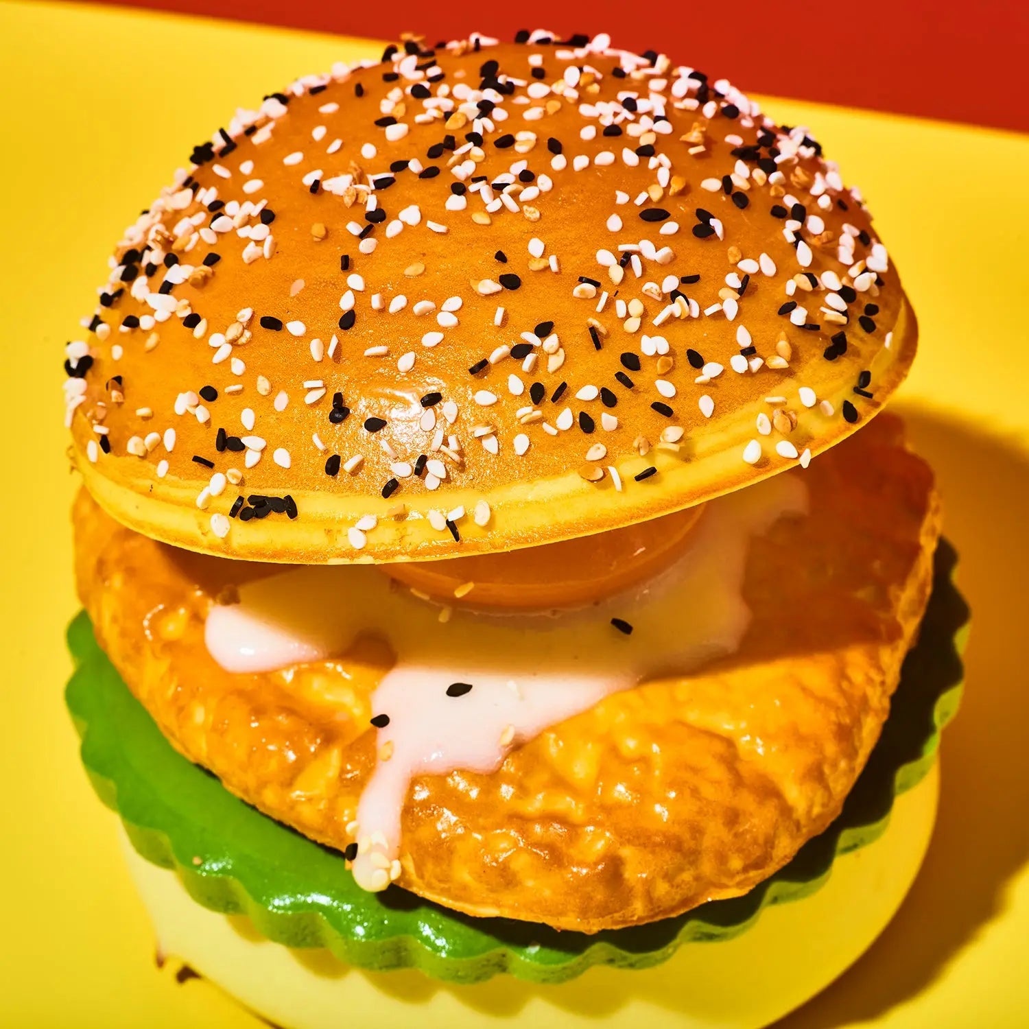 A close-up of a novelty breakfast sandwich toy or prop on a bright yellow background. The sandwich features a golden-brown waffle top sprinkled with black and white sesame seeds, a textured golden-brown patty with dripping white sauce, a green fluted bottom, and a fried egg in the center.